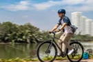 A man with a blue helmet riding a grey commuter bike along a scenic multi-use waterfront path, with city high-rises and a lake in the background, illustrating the practical benefits and savings from buying a used bike.