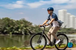 A man with a blue helmet riding a grey commuter bike along a scenic multi-use waterfront path, with city high-rises and a lake in the background, illustrating the practical benefits and savings from buying a used bike.