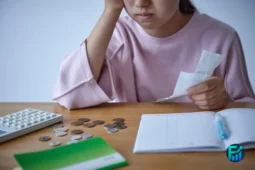 A worried person looking at a bill with scattered coins and a calculator on a table, highlighting the need for emergency cash assistance.